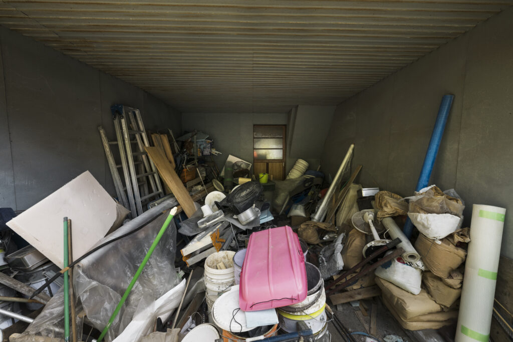 A cluttered storage room filled with boxes, bags, tools, and construction materials, including a pink toolbox and metal shelves.