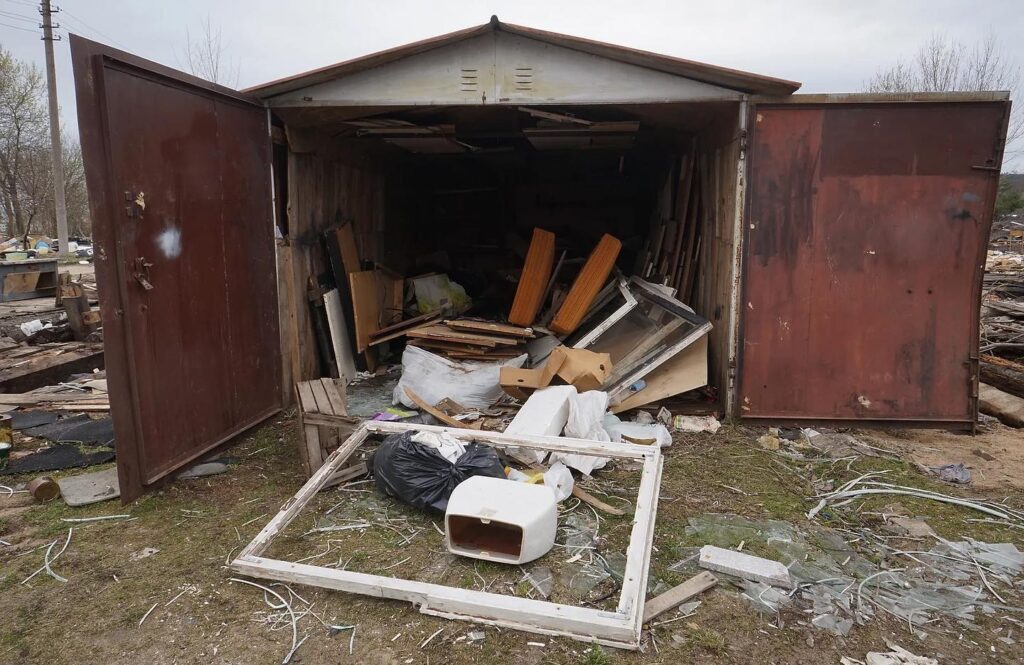 Interior of a large rusted shipping container used as a storage shed, filled with scattered construction materials, boards, metal pieces, and debris on the ground, Furniture Removal