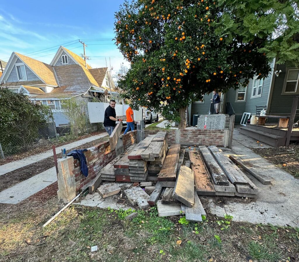 Man carrying construction materials near a brick and stone project site, with an orange fruit tree in the background.