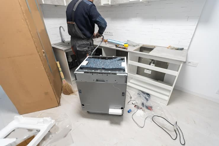 A handyman installing a dishwasher in a kitchen with white brick-patterned tile walls and white cabinetry. The dishwasher is positioned in the center, with the worker behind it adjusting the countertop area. A large cardboard box sits on the floor to the left.
