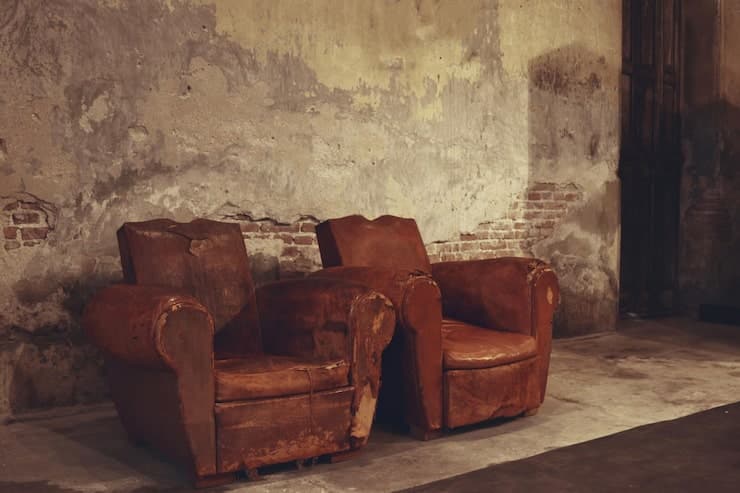 Two worn brown leather armchairs sit side by side in a decaying room with peeling paint that exposes brickwork beneath. Dim lighting casts shadows across the chairs' torn upholstery and faded surfaces, giving the scene an atmosphere of abandonment and age.