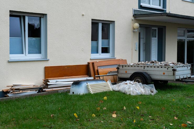 Cream-colored building with windows and blinds, renovation debris and a utility trailer piled with rubble on its lawn, beside a plastic-wrapped object and a discarded white bathtub.