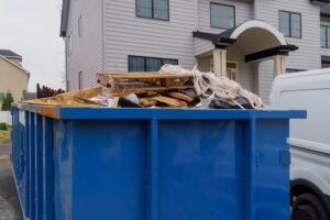 A large blue dumpster overflowing with construction debris—wooden pallets, planks, and plastic sheeting—sits in the driveway of a modern two-story house with light grey siding.