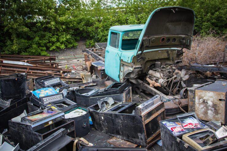 A junkyard scene shows a stripped-down light blue pickup truck with its hood open revealing the engine, surrounded by haphazardly piled black containers from old arcade games and slot machines, with rusty metal beams and lush green foliage in the background.