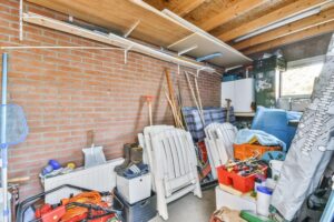 A cluttered garage storage room with brick walls and wooden beams, filled with shelves, tools, pool equipment, Adirondack chairs, bins, and boxes.